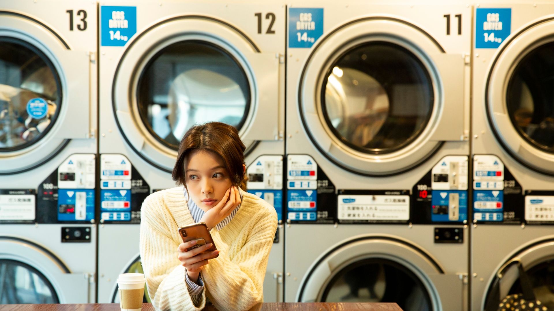 Person waiting at laundromat, looking at phone with coffee nearby