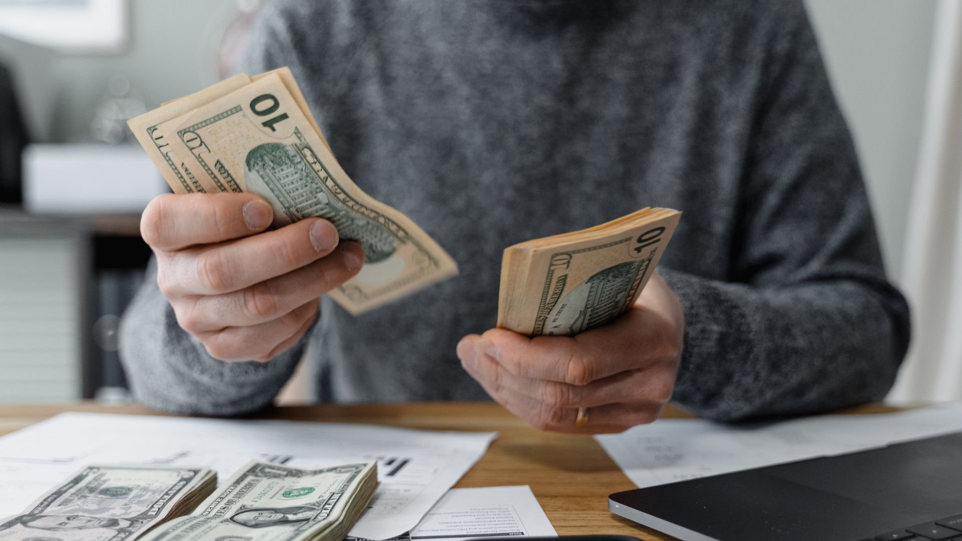 Person in gray sweater counting ten-dollar bills at a desk